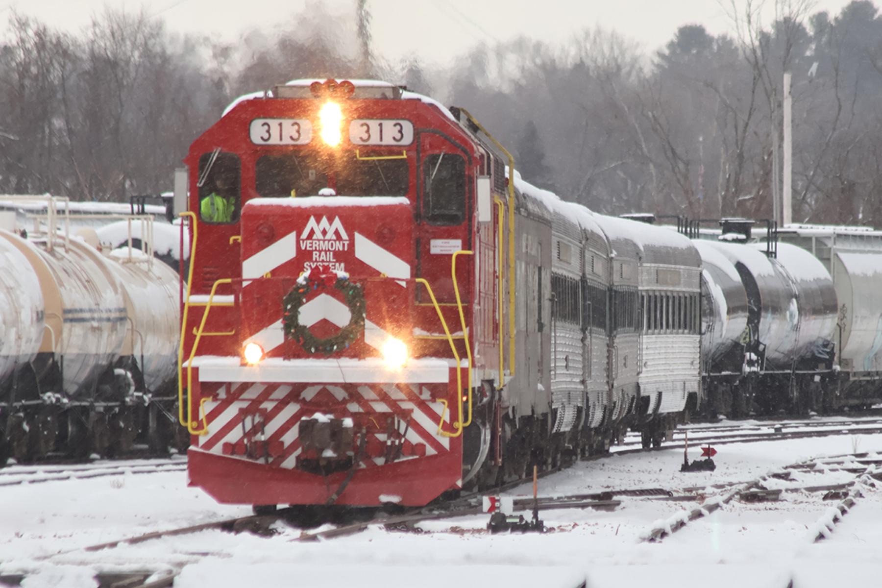 The Polar Express train arriving through falling snow, its engine decorated with a holiday wreath